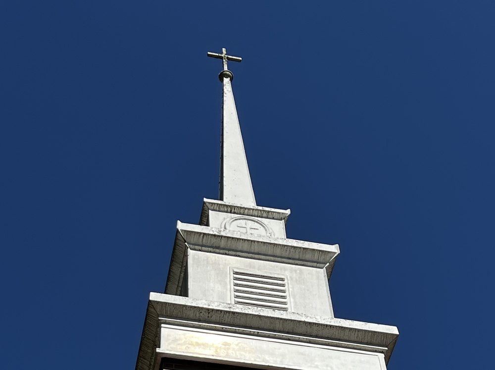 A white church steeple and cross rise above a red brick church against a clear blue sky.